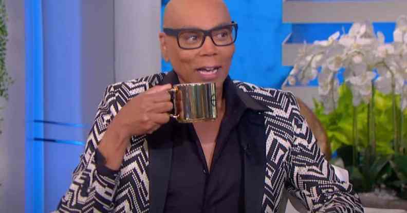 RuPaul appears on the Ellen DeGeneres show in a black, white and grey patterned suit, holding a gold coloured mug
