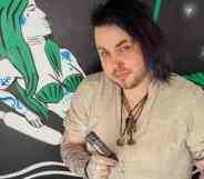 Alden Tank is dressed in a tan top and holds a tattoo gun standing in front of a green mermaid/siren painted onto a wall