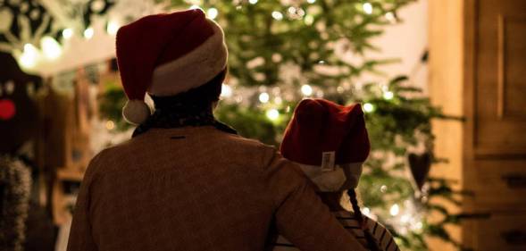 an older person wraps their arm around a younger person. Both are wearing Christmas hats as they look on at a Christmas tree