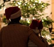 an older person wraps their arm around a younger person. Both are wearing Christmas hats as they look on at a Christmas tree
