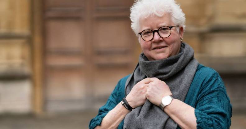Val McDermid, a white woman, wears a blue shirt and a grey scarf that her hands are clutching onto