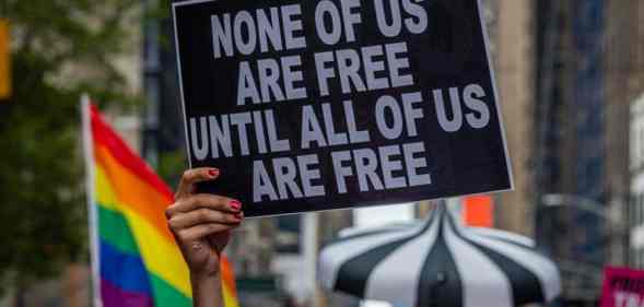 A participant holds a sign reading "None of us are free until all of us are free" and an LGBT+ Pride flag can be seen behind during the Reclaim Pride Coalition's third annual Queer Liberation March