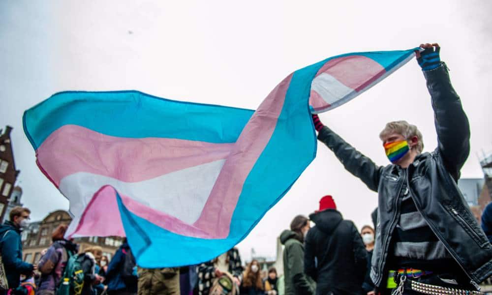 A person wearing a rainbow coloured face mask holds up a transgender Pride flag above their head