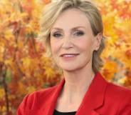 Jane Lynch, a white woman with blonde hair, wears a black shirt and red jacket while standing in front of orange leaves
