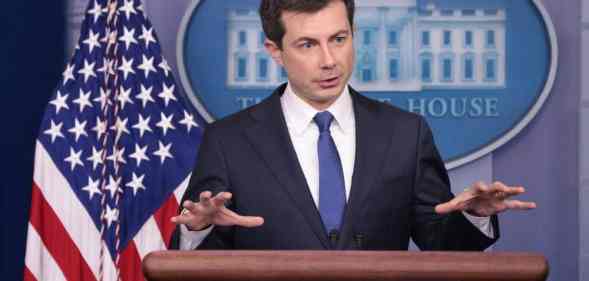 Pete Buttigieg stands at a podium dressed in a suit and tie with his hands raised