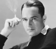 William Haines, an American actor, wears a white shirt and a dark sweater on top while holding a cigarette