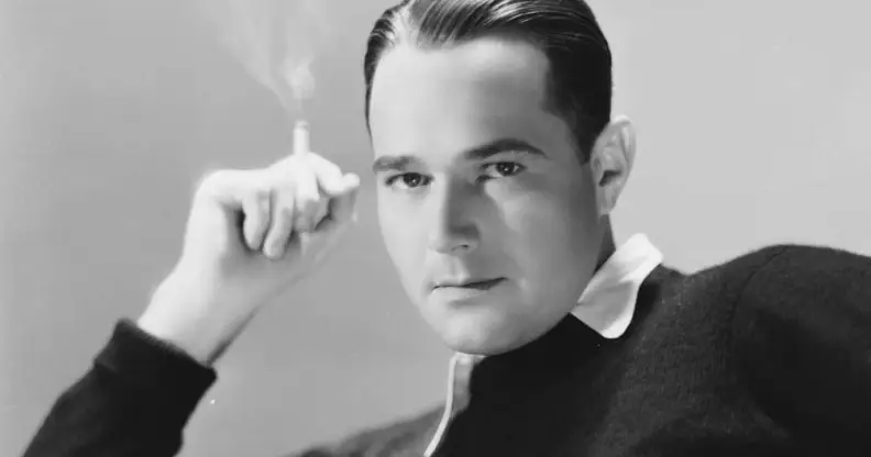 William Haines, an American actor, wears a white shirt and a dark sweater on top while holding a cigarette