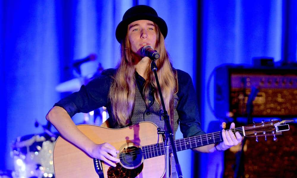 Sawyer Fredericks wears a bowler hat, grey vest and black long sleeved shirt as he sings and plays the guitar on stage