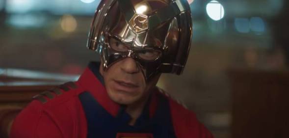 Peacemaker, played by John Cena, looks incredulously at other people gathered around a table