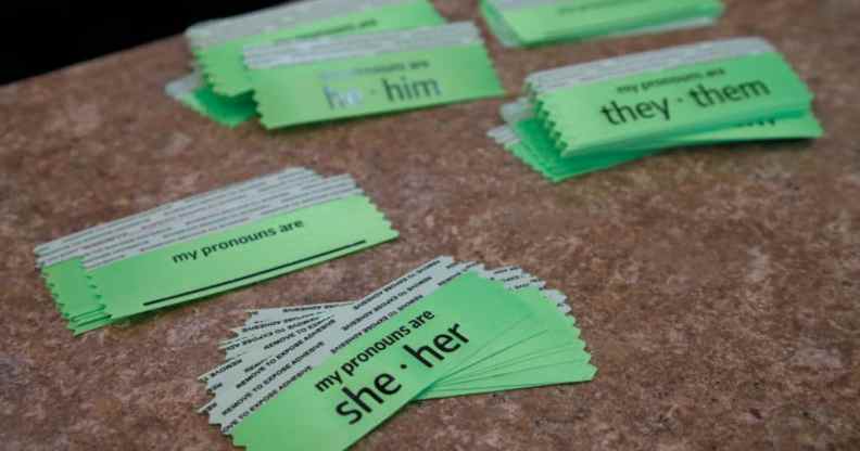 Pronoun stickers on a table, reading 'she/her', 'he/him', and 'they/them' along with blank ones