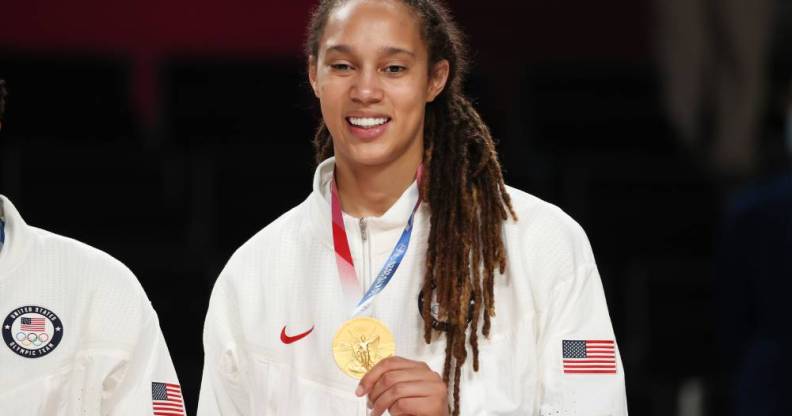 Brittney Griner, a basketball player, holds up her Olympic gold medal while wearing a white Team USA jacket