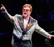 Elton John wears a patterned and sparkly jacket with red tinted glasses and holds his arms up during a performance