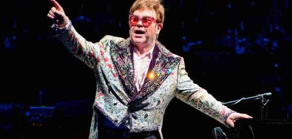 Elton John wears a patterned and sparkly jacket with red tinted glasses and holds his arms up during a performance