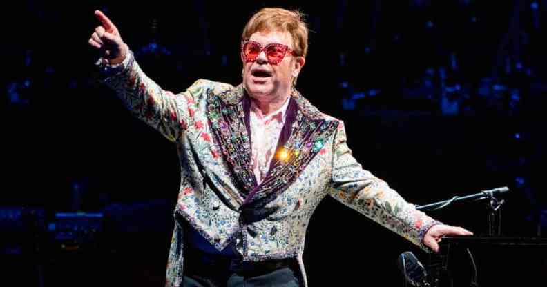 Elton John wears a patterned and sparkly jacket with red tinted glasses and holds his arms up during a performance