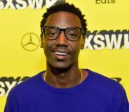 Jerrod Carmichael smiles at the camera while wearing a blue-purple top in front of a bright yellow background