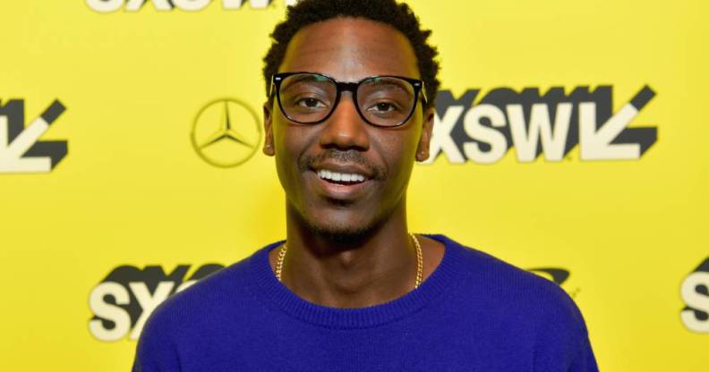 Jerrod Carmichael smiles at the camera while wearing a blue-purple top in front of a bright yellow background