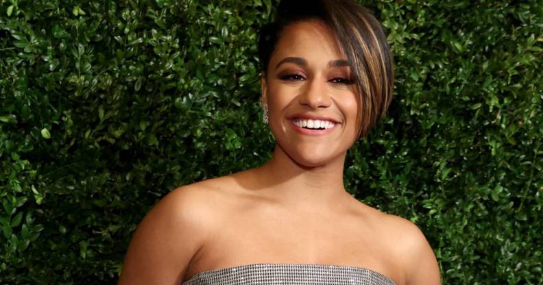 Ariana DeBose smiles at the camera while standing in front of a background filled with greenery