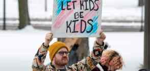 A person holds up a sign reading "let kids be kids" in front of the colours of the transgender pride flag