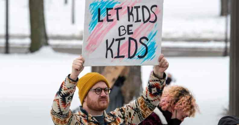 A person holds up a sign reading "let kids be kids" in front of the colours of the transgender pride flag
