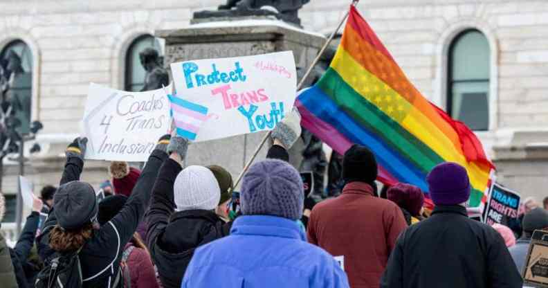A group of people gather together to protest on behalf of the trans community with a sign reading "protect trans youth" visible in the picture