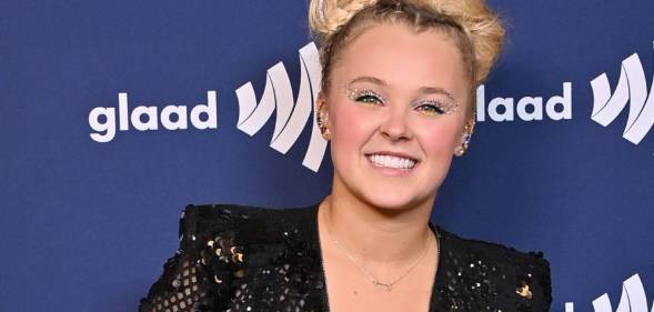 JoJo Siwa smiles at the camera as she wears a black sequinned outfit and stands in front of a dark blue background with the white GLAAD logo on it