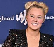 JoJo Siwa smiles at the camera as she wears a black sequinned outfit and stands in front of a dark blue background with the white GLAAD logo on it