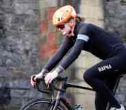 Trans cyclist Emily Bridges rides a black bicycle in front of a brick building