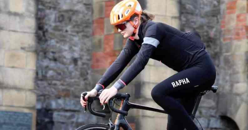 Trans cyclist Emily Bridges rides a black bicycle in front of a brick building
