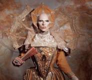 Cheddar Gorgeous, a drag artist, is dressed in a gold elizabethan gown with a matching headdress and hand fan