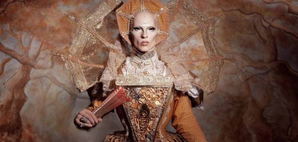 Cheddar Gorgeous, a drag artist, is dressed in a gold elizabethan gown with a matching headdress and hand fan