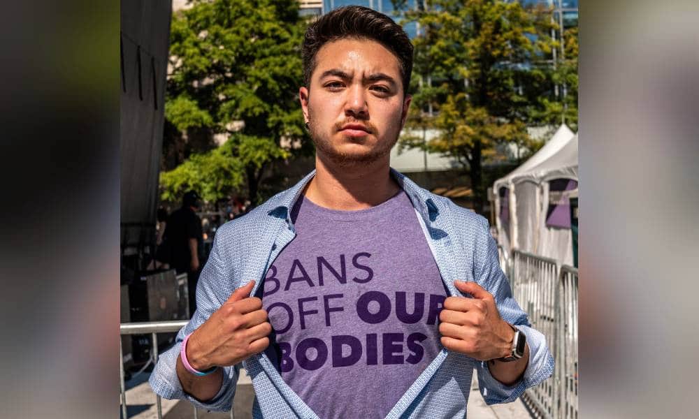 Schuyler Bailar holds open a blue button up shirt while wearing a purple shirt underneath with the worlds 'bans off our bodies' on it