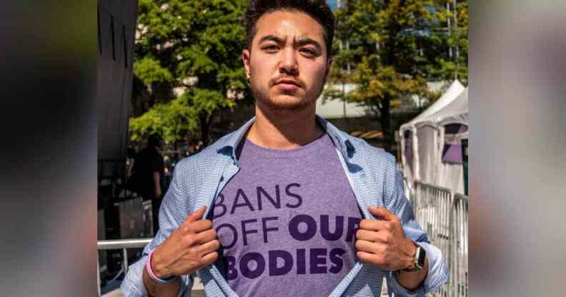 Schuyler Bailar holds open a blue button up shirt while wearing a purple shirt underneath with the worlds 'bans off our bodies' on it