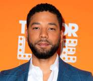 Jussie Smollett stares at the camera while wearing a white shirt and blue jacket as he stands in front of an orange background