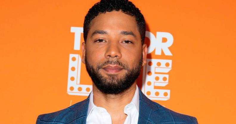 Jussie Smollett stares at the camera while wearing a white shirt and blue jacket as he stands in front of an orange background
