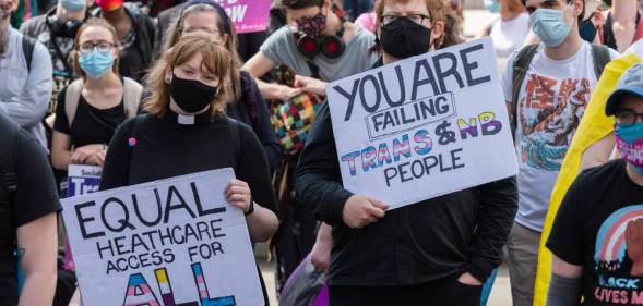 Two people hold up signs during a protest. One sign reads 'You are failing trans and NB [non-binary] people' and the other sign reads 'equal healthcare access for all'