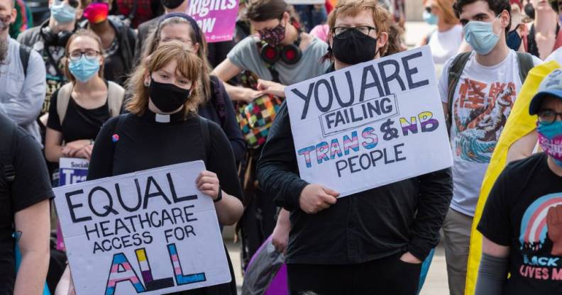 Two people hold up signs during a protest. One sign reads 'You are failing trans and NB [non-binary] people' and the other sign reads 'equal healthcare access for all'