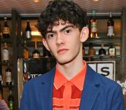 Heartstopper star Joe Locke stares at the camera while wearing an orange patterned shirt and blue jacket