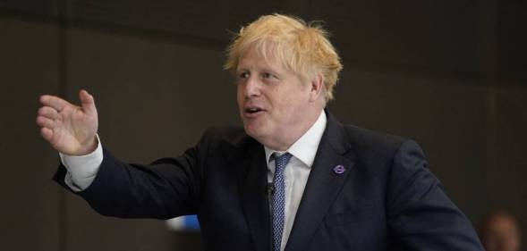 Boris Johnson wears a dark suit jacket, white shirt and blue tie as he gestures to the side