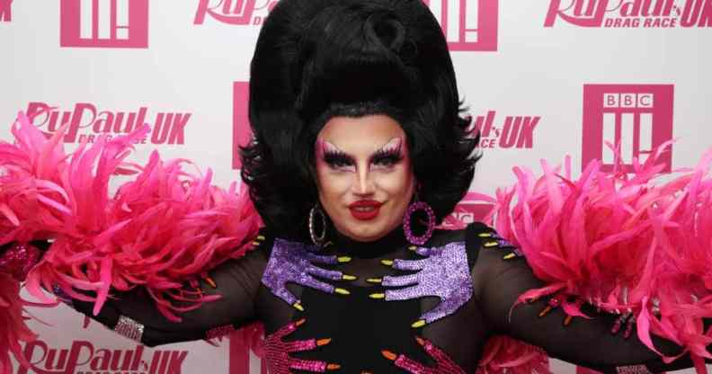 Drag Race UK star Choriza May stares at the camera while wearing a black wig, black outfit with pink and purple hands on it