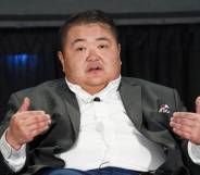 Emmett Schelling sits in a chair while wearing a white button up shirt and grey jacket. He is gesturing with both hands towards himself