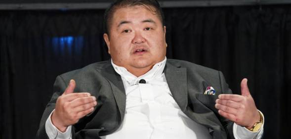 Emmett Schelling sits in a chair while wearing a white button up shirt and grey jacket. He is gesturing with both hands towards himself