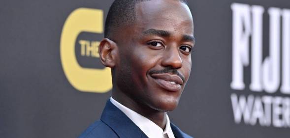 Ncuti Gatwa smiles at the camera while wearing a white shirt and dark blue coat