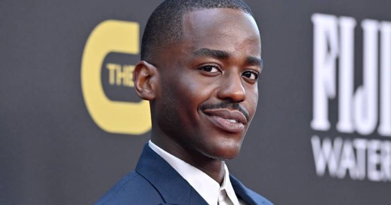 Ncuti Gatwa smiles at the camera while wearing a white shirt and dark blue coat