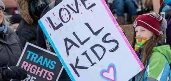 A young person holds up a sign that reads 'Love all kids' while a sign reading 'trans rights are human rights' is in the background