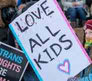 A young person holds up a sign that reads 'Love all kids' while a sign reading 'trans rights are human rights' is in the background