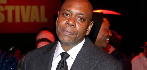 Dave Chappelle stares at the camera while wearing a black tie, white shirt and dark jacket