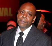 Dave Chappelle stares at the camera while wearing a black tie, white shirt and dark jacket