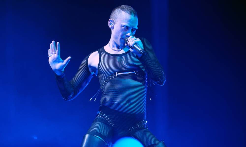 Years & Years singer Olly Alexander sings into a microphone while wearing a black mesh bodysuit and being lit by a blue light