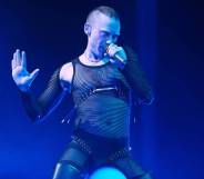Years & Years singer Olly Alexander sings into a microphone while wearing a black mesh bodysuit and being lit by a blue light