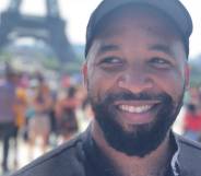 Michael Reynolds, a Black LGBTQ+ gamer with a beard, smiles as he stares off screen. He is wearing a black shirt and black cap as he stands outside